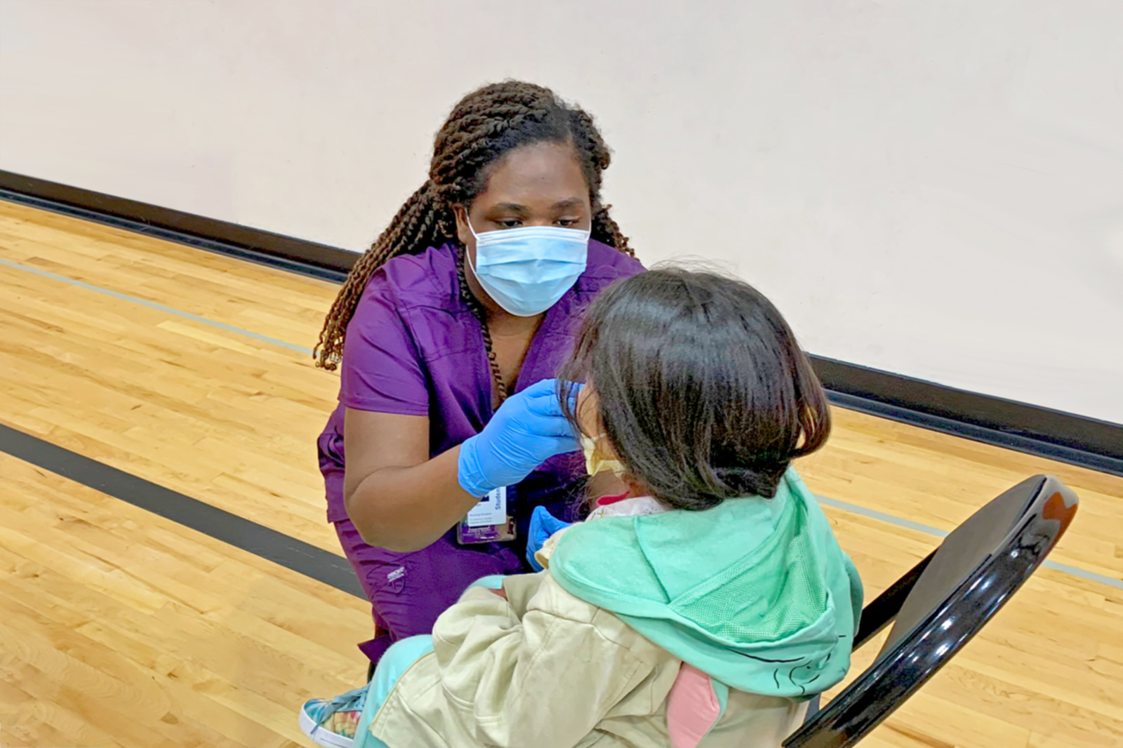 Katilyn Taylor MSN-EL'22, in purple scrubs, kneels down in front of a student to administer a flu vaccine during her clinical experience at the Bear Care Health and Wellness Clinic in fall 2020.
