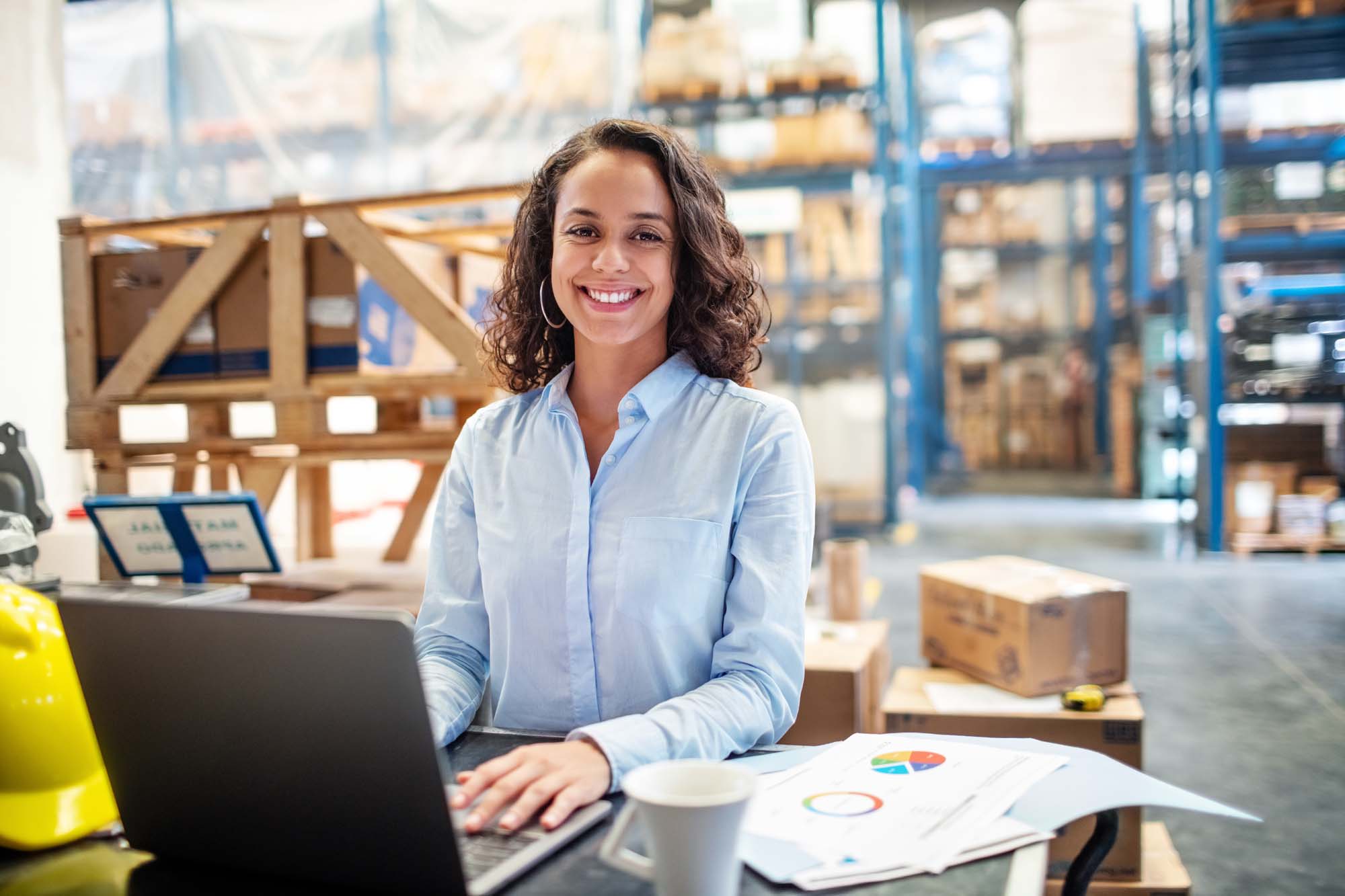 A smiling Business Management graduate, working on a laptop at a desk inside a warehouse