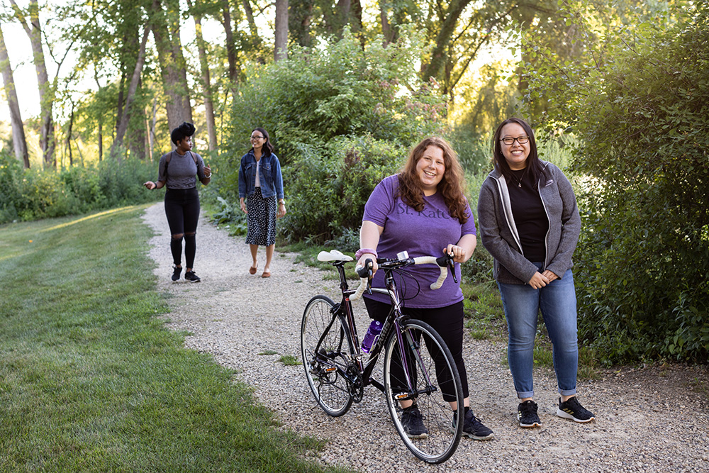 Four students walking on a trail in the woods. One is walking a bicycle.