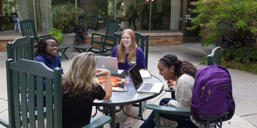 Four students gathered around outdoor table, sharing pizza and laughing.