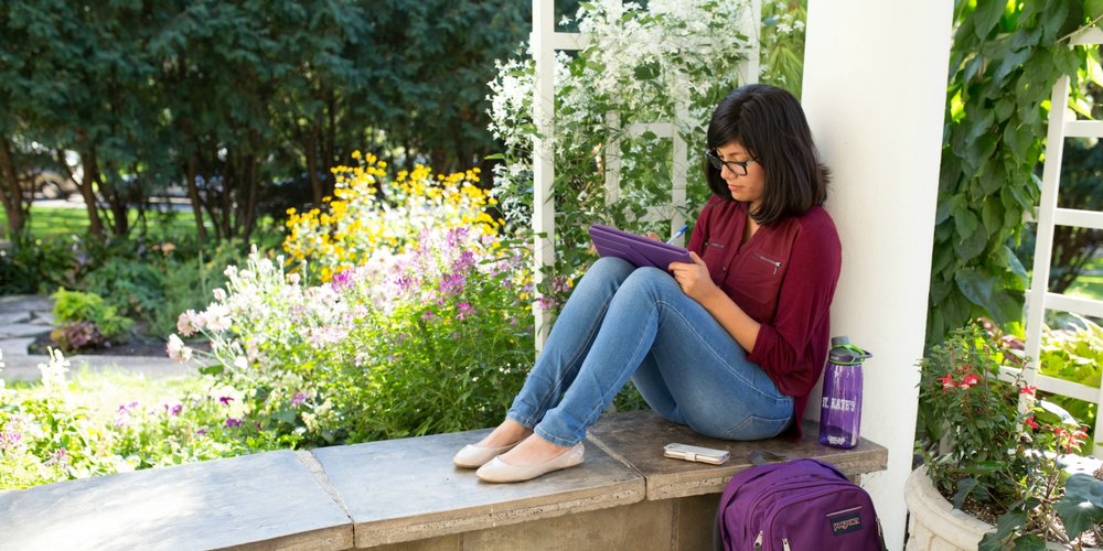 Student sitting in a campus common area, holding a laptop with books and backpacks nearby
