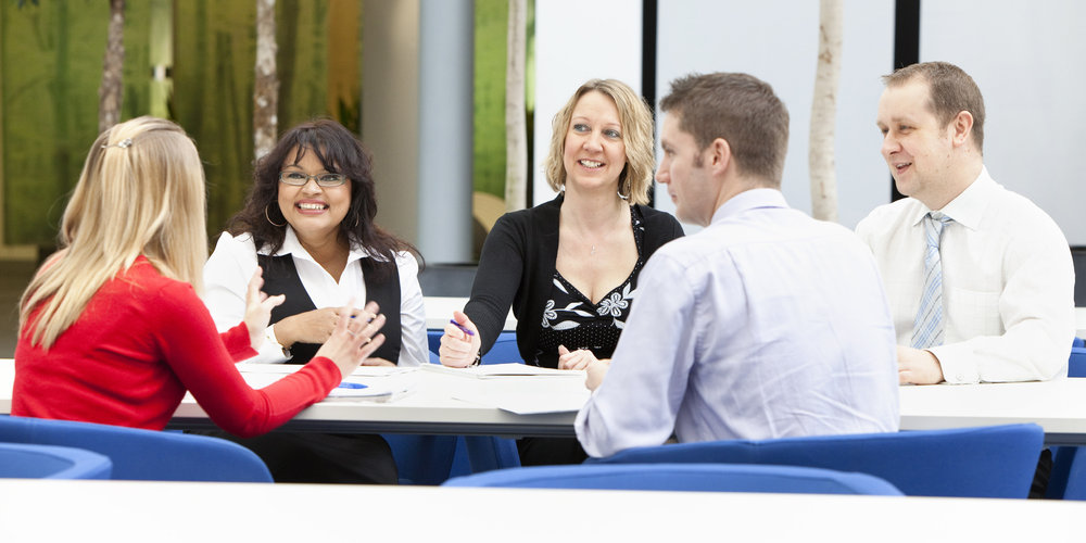 Five graduate students engaged in a collaborative work meeting around a table, discussing documents and using a laptop