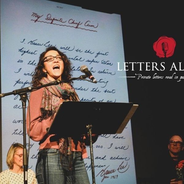 Woman standing at a microphone with a large handwritten letter behind her