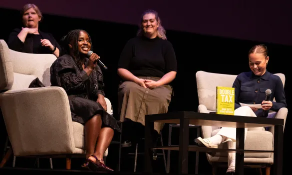 Anna Gifty Opoku-Agyeman sits on stage and speaks into a microphone. Sitting across from her is Hope Walz and behind them sit two sign language interpreters.