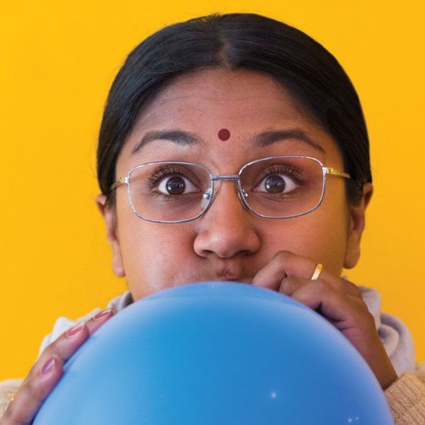Person with glasses and a red bindi blowing up a blue balloon, against a bright yellow background.